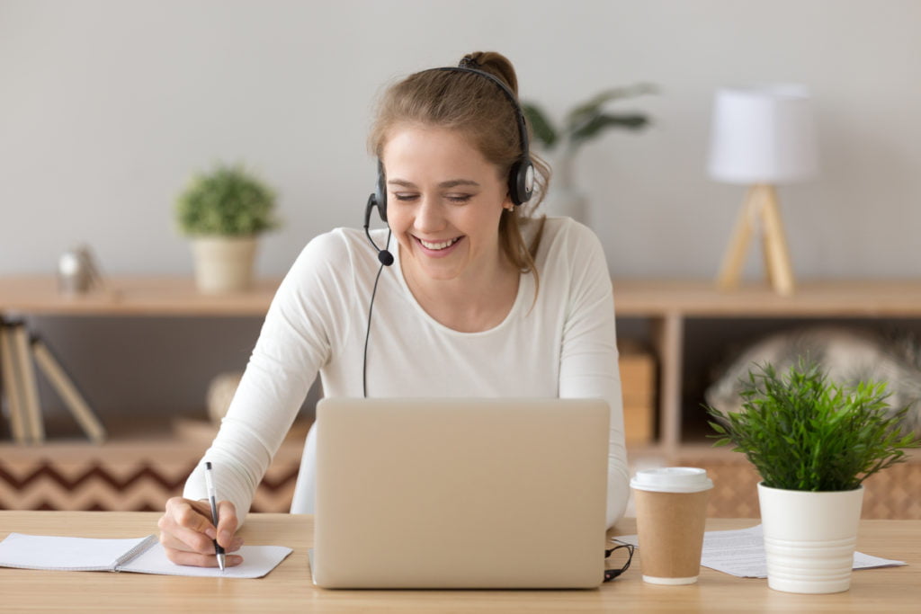 Smiling woman wearing headset writing notes studying online on laptop